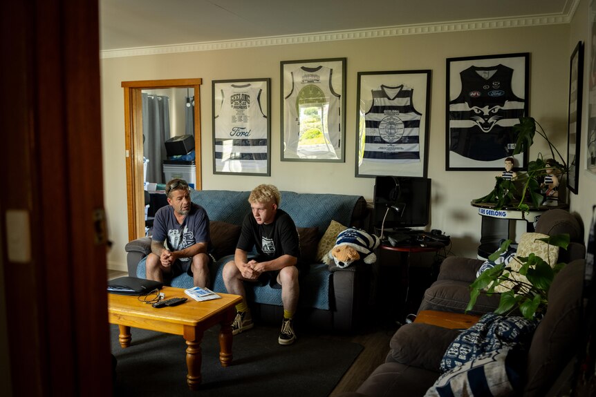 Teenage boy sits on couch next to dad in living room with framed Geelong Cats jerseys framed on the wall behind them.