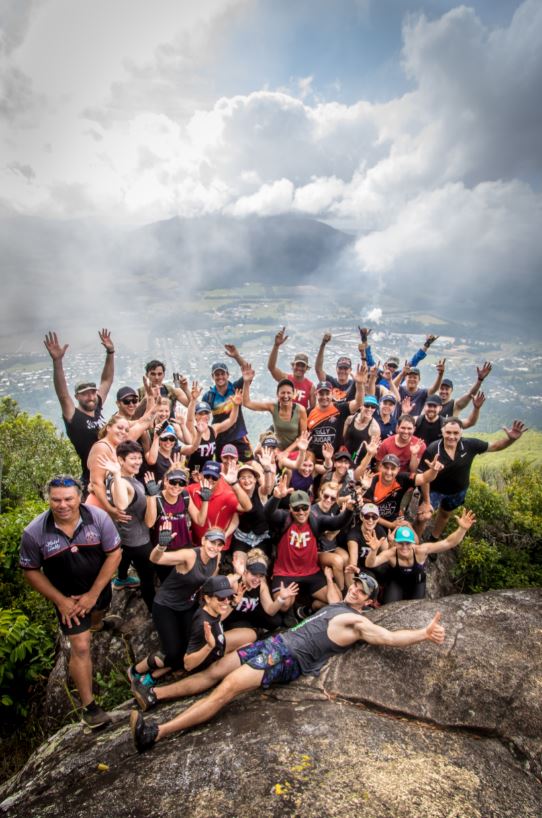 A group of people smile and wave as they pose at the top of a mountain.