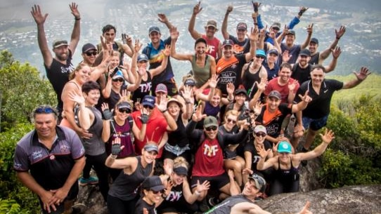 A group of people smile and wave as they pose at the top of a mountain.
