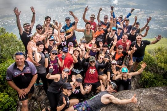 A group of people smile and wave as they pose at the top of a mountain.