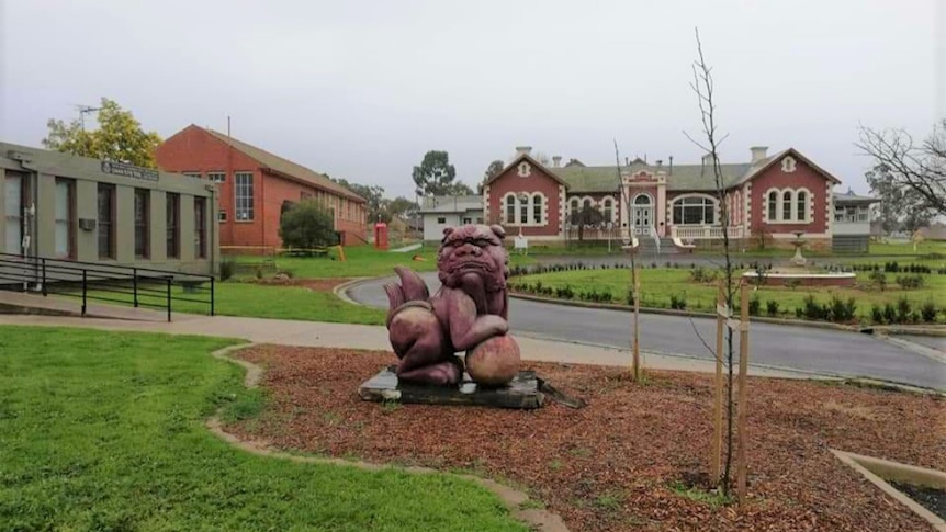 A gargoyle, head facing the camera, sits in a garden bed. Behind it is a gothic burgundy-coloured building 