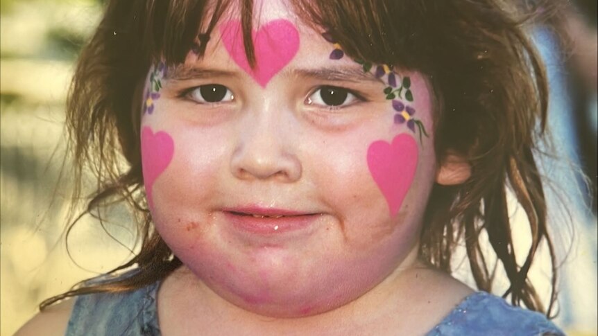 A young girl with brunette hair and pink face paint stares into the camera.