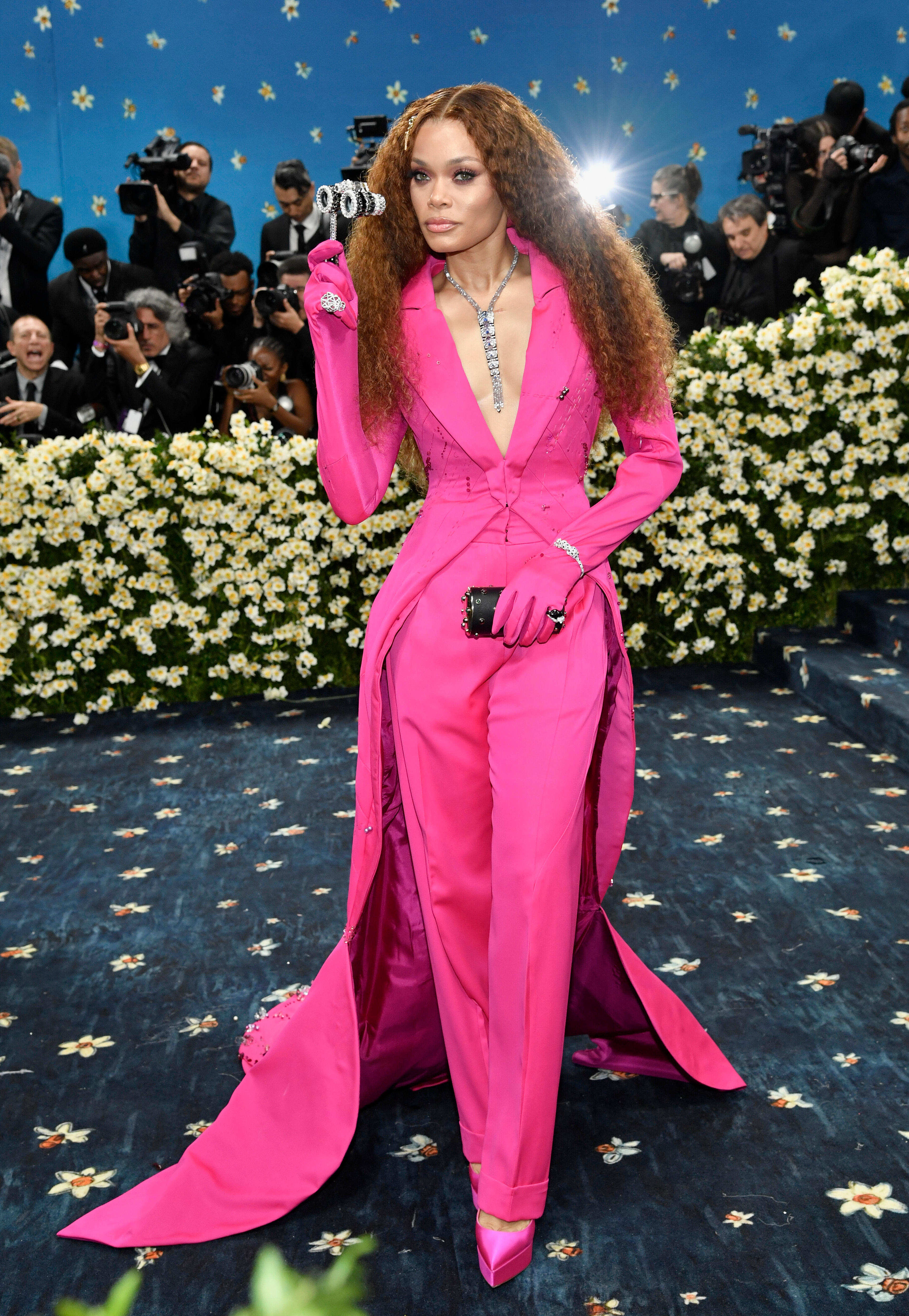 Andra Day wearing a pink pantsuit with matching gloves, holding a set of bejewelled opera binoculars 