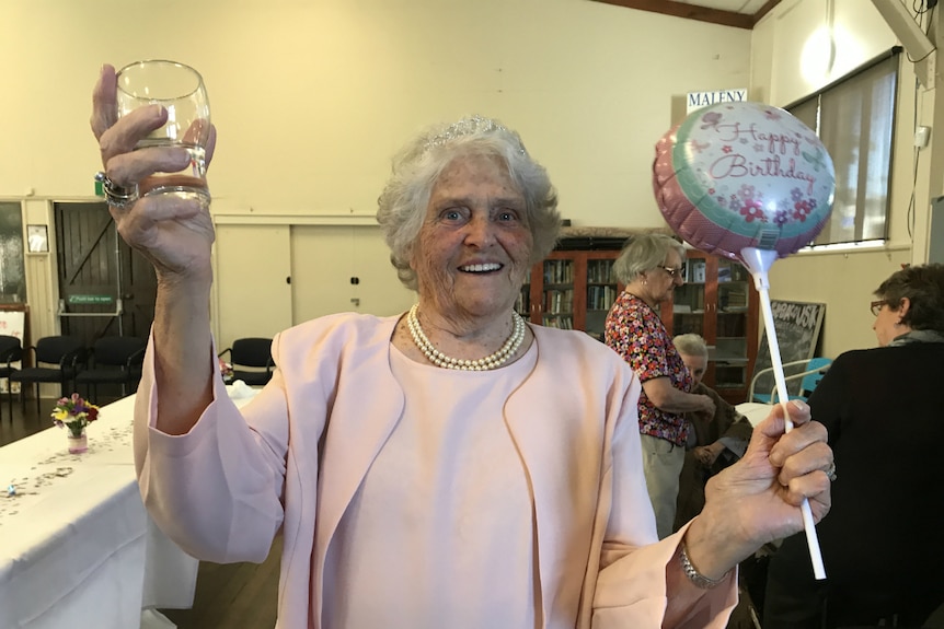 Mrs Woods holds up a glass of lemonade in one hand, smiling at the camera with a happy birthday balloon in her other hand.