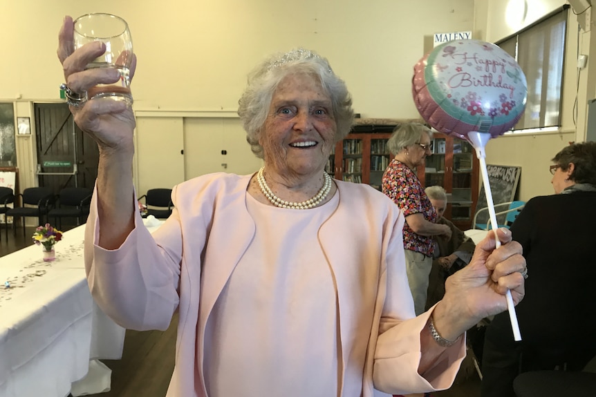 Mrs Woods holds up a glass of lemonade in one hand, smiling at the camera with a happy birthday balloon in her other hand.