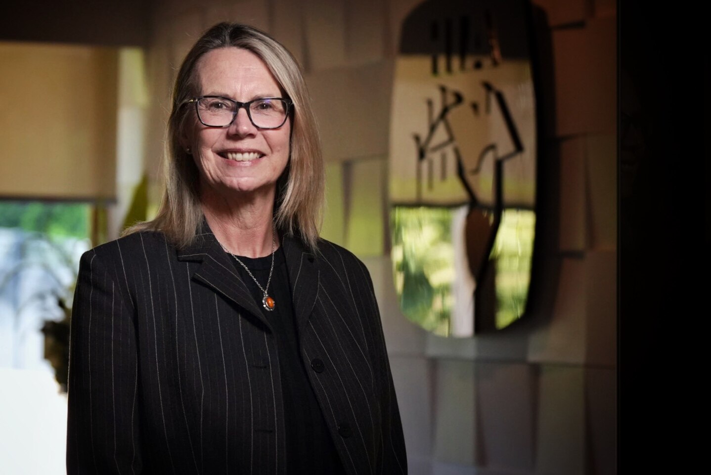 Smiling Jocelyn has blond shoulder-length hair, wears glasses, pinstripe black jacket, top, room with mirror behind.
