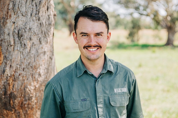 A man in his thirties with dark hair and a moustache stands smiling in front of a camera, with a tree and paddock behind him.