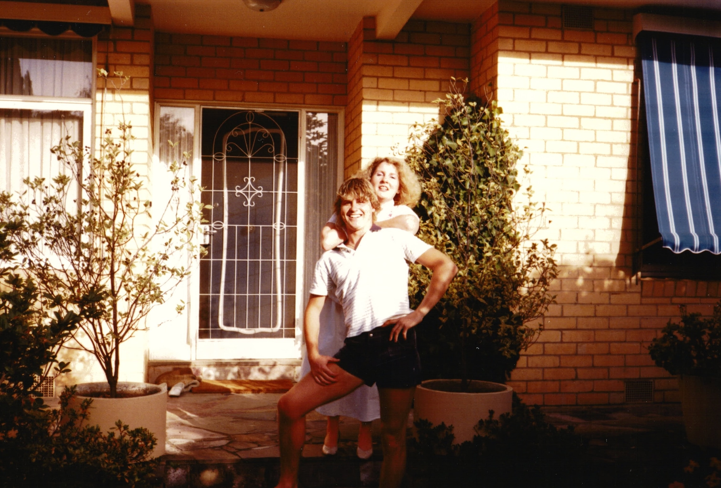 A young man and woman smile as they pose in a comical way at the entrance to a home.