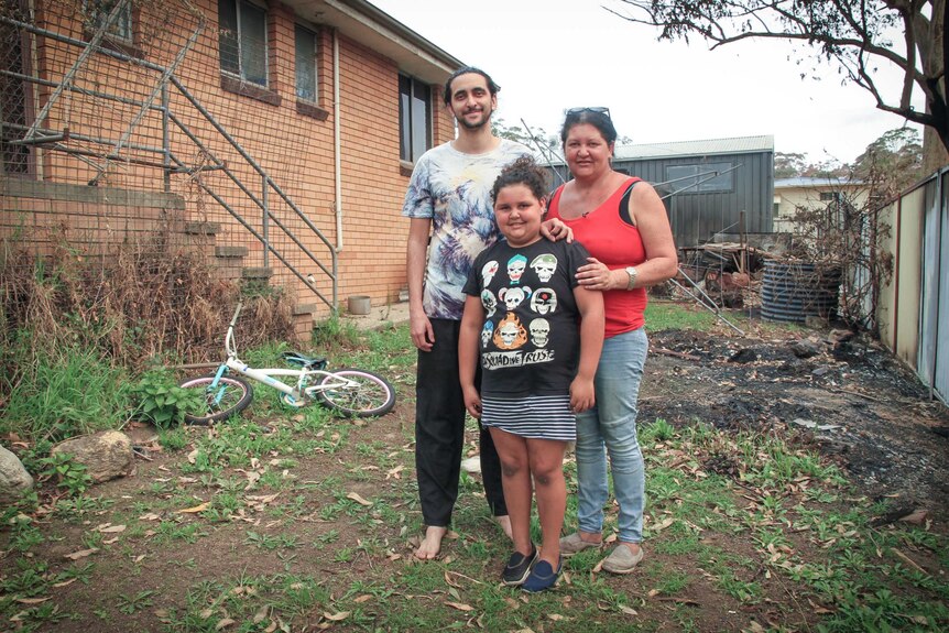 Mother with adult son and young daughter standing beside house with burnt yard and fence.