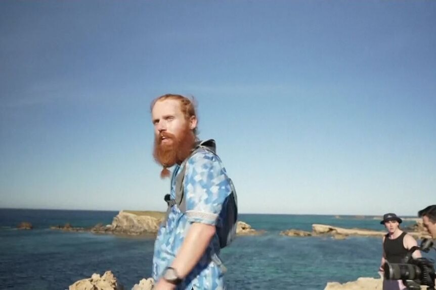 Red bearded man wearing runner backpack and blue plaid shirt looks off edge of cliff in Tunisia. 