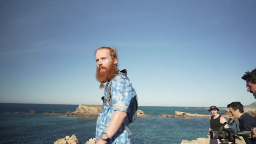 Red bearded man wearing runner backpack and blue plaid shirt looks off edge of cliff in Tunisia. 