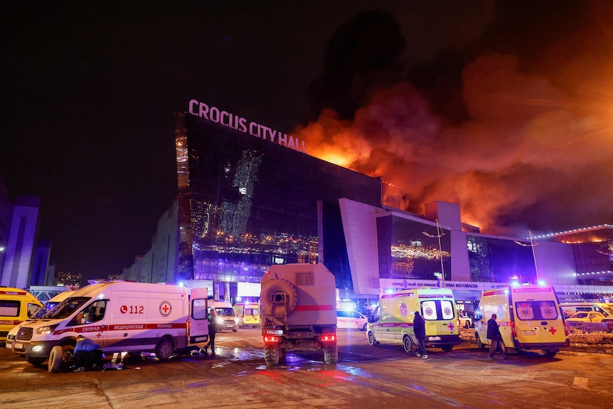 Rows of ambulances parked outside concert hall with flames and smoke emerging from the roof.