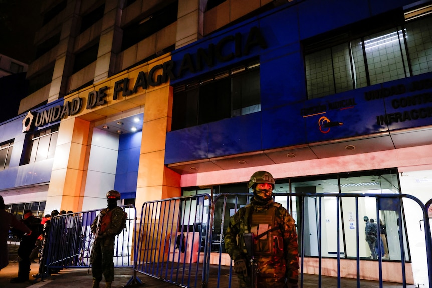 Heavily armed soldiers in camouflage uniforms stand outside a blue and yello building at night.