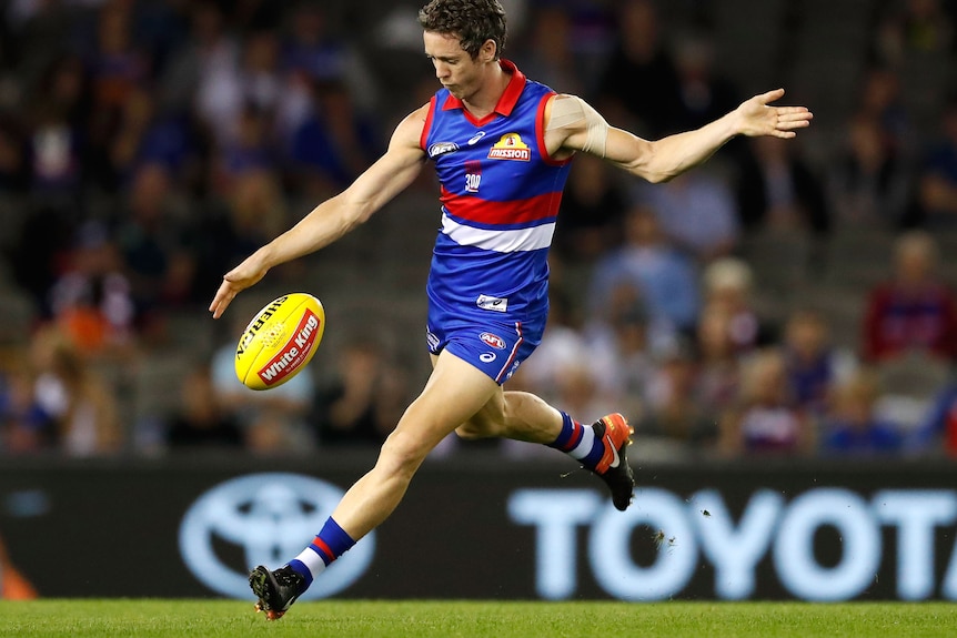 A male AFL player in a blue and red uniform captured mid kick. He is dropping the ball in front of him and his leg is extended.
