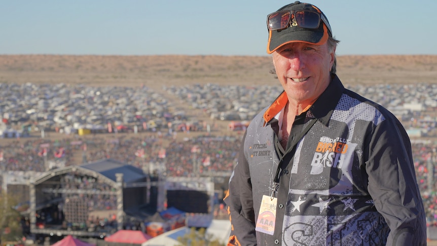 A man in a cap stands on a sandune hill with a big festival crowd behind him