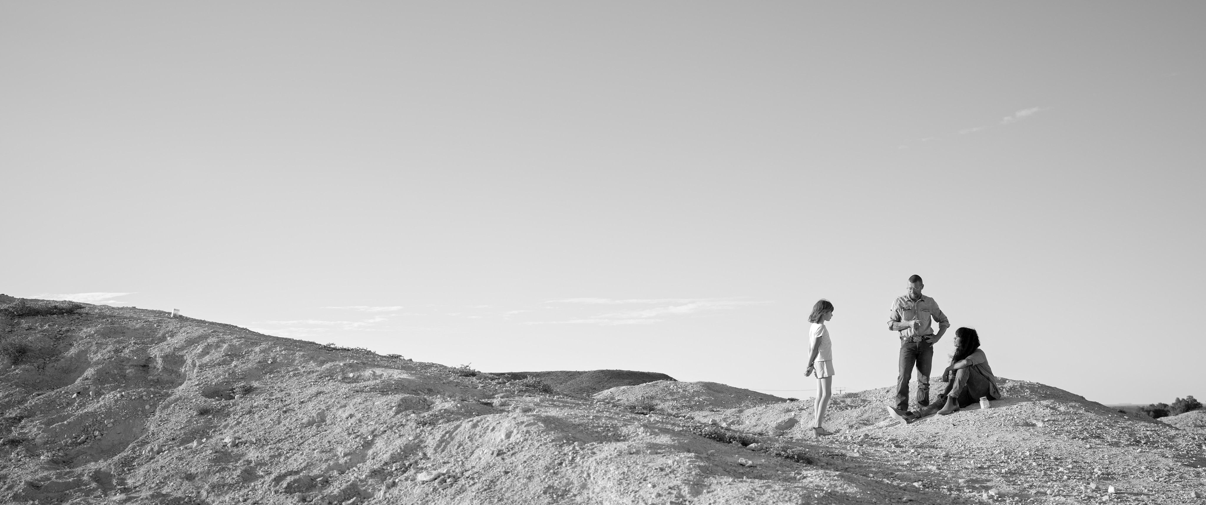 A man stands and talks to a woman and a girl in a vast outback setting. The woman is seated on the dirt.