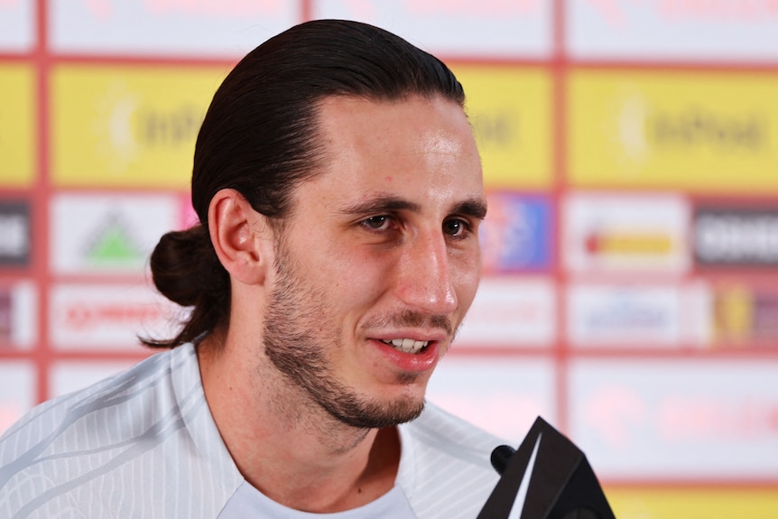 A young man with long hair and beard smiles as he speaks into press conference microphone