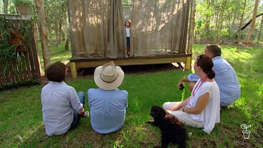 Adults and a dog sitting around outdoor stage in a garden with children performing