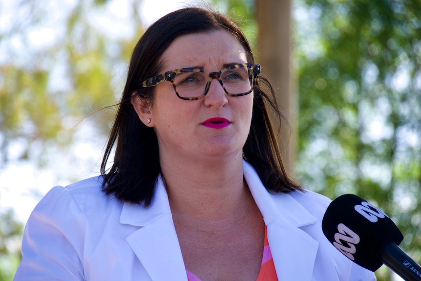 A woman with brown hair and glasses, wearing a orange shirt and a white jacket. 