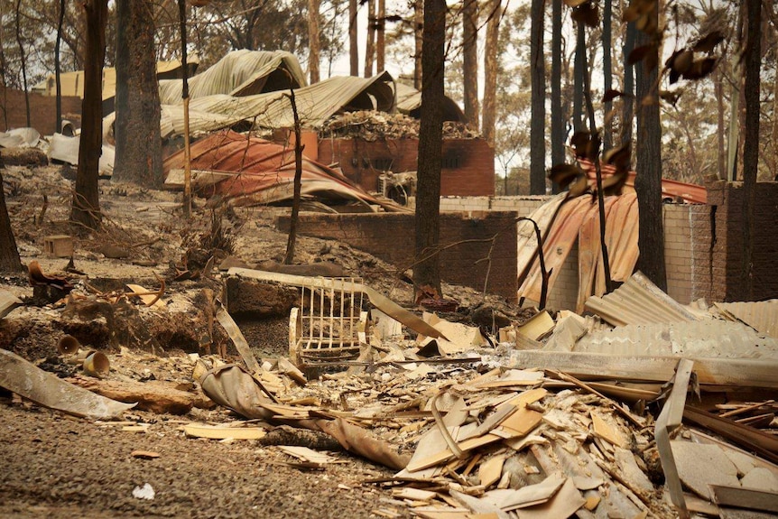 A chair sits upright among rubble and warped iron left, with blackened bushland visible nearby.