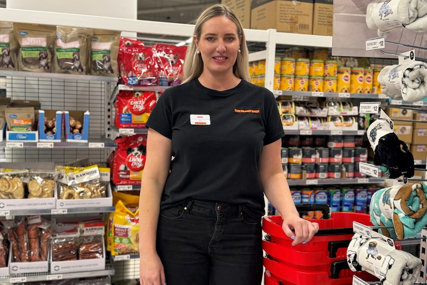 A woman with long blonde hair wearing a black t-shirt and black jeans stands in front of an aisle of pet food in a store.
