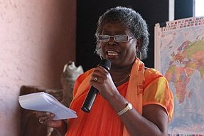 A woman speaking into a microphone and holding a notepad standing in front of a map of the world