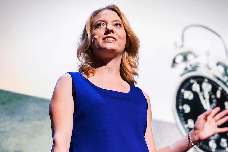 Woman with shoulder-length blonde hair and bright blue top stands looking to the side, wearing a microphone headset.