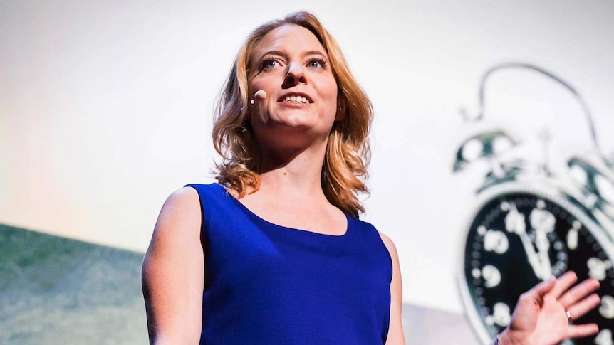 Woman with shoulder-length blonde hair and bright blue top stands looking to the side, wearing a microphone headset.