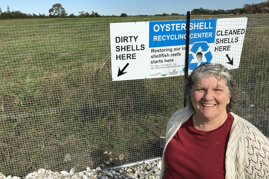 A woman in a burgundy t-shirt and cream cardigan stands in front of a recycling sign, with a paddock in the background.