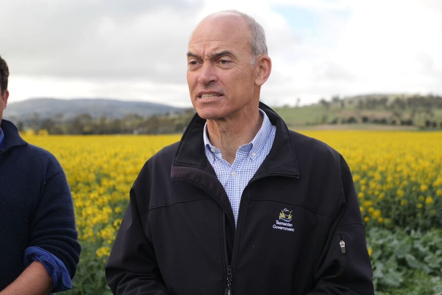 A bald man in a government jacket and blue shirt stands in front of a yellow field. 