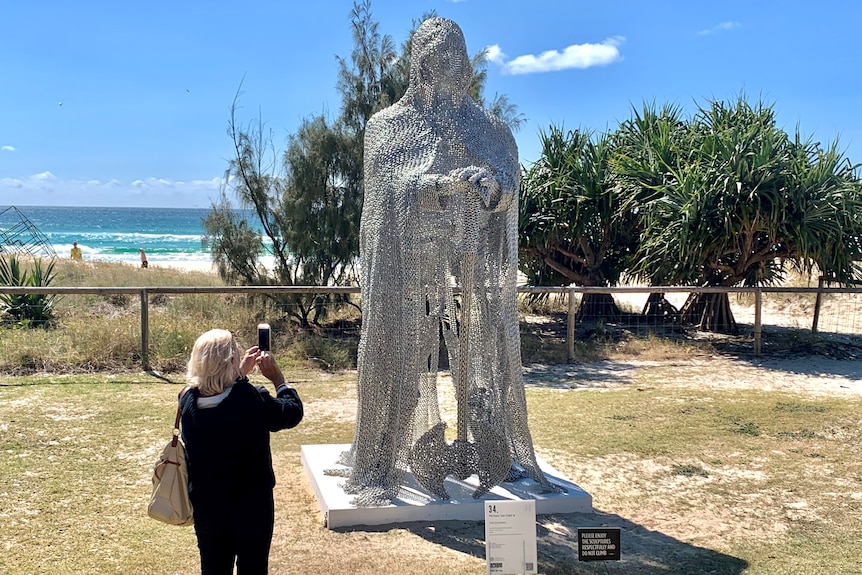 Sculpture of mesh guardian figure towering over person taking a photo at Currumbin beach Swell sculpture festival
