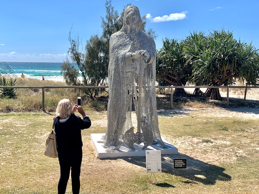 Sculpture of mesh guardian figure towering over person taking a photo at Currumbin beach Swell sculpture festival