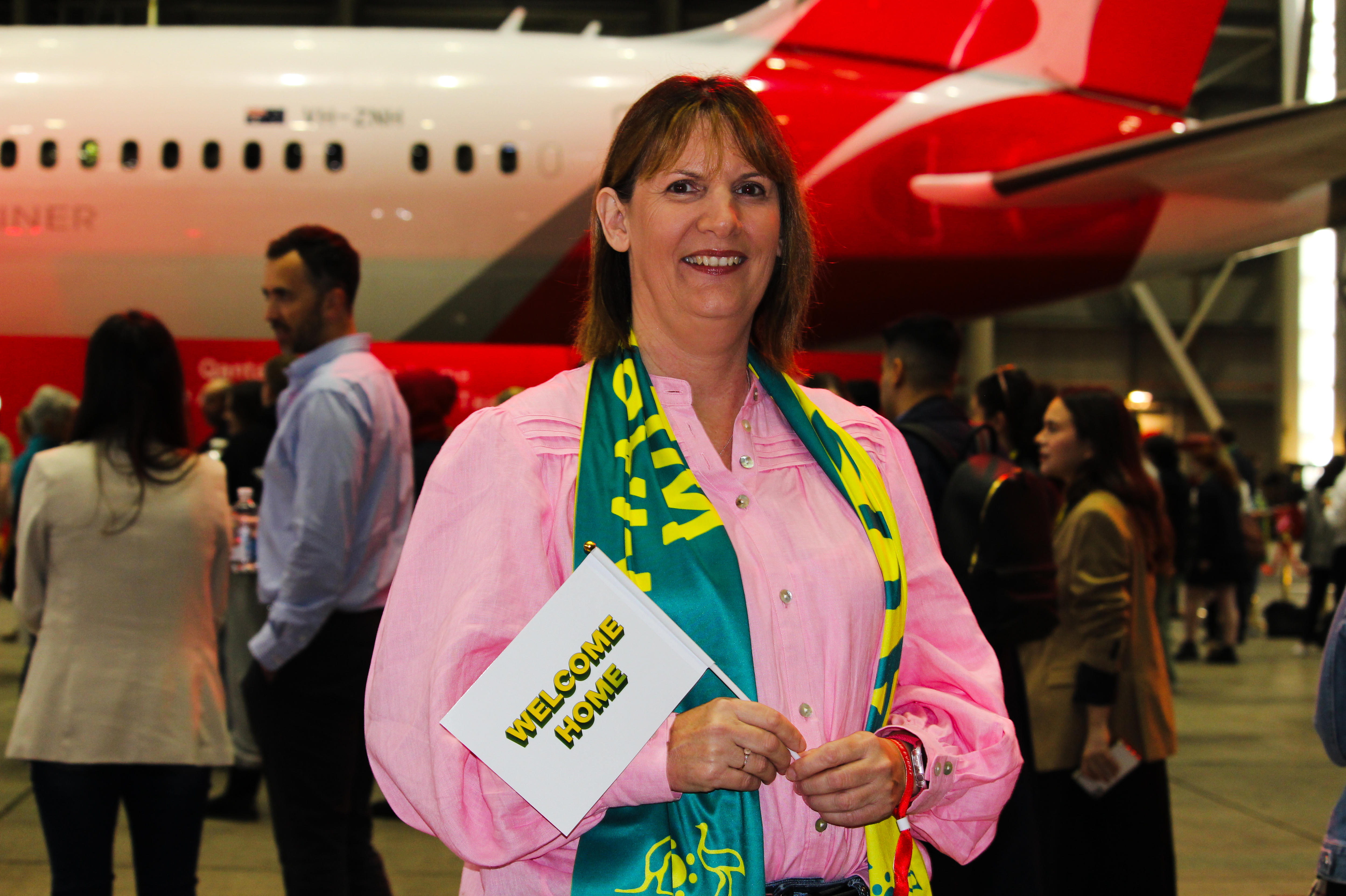 A woman wearing a green and gold scarf and a sign that reads WELCOME HOME standing in front of a Qantas plane