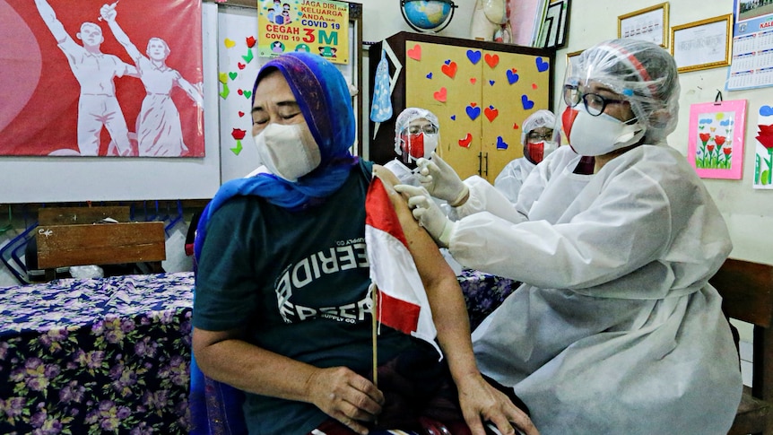 A woman in a hijab receives an injection from a nurse in full PPE.