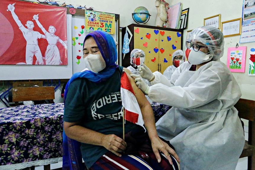 A woman in a hijab receives an injection from a nurse in full PPE.