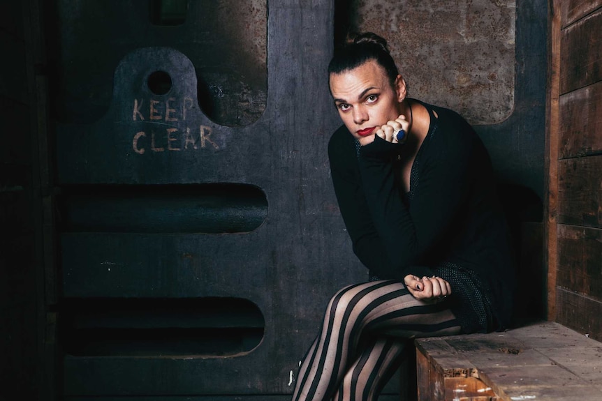 Cassie Workman, wearing a black top and striped pants, sits against a rusted metal background.