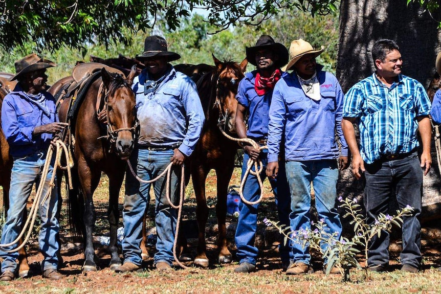 A group of five stockmen stand in front of their horses