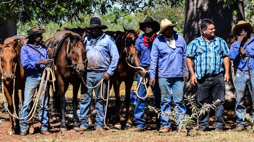 A group of five stockmen stand in front of their horses