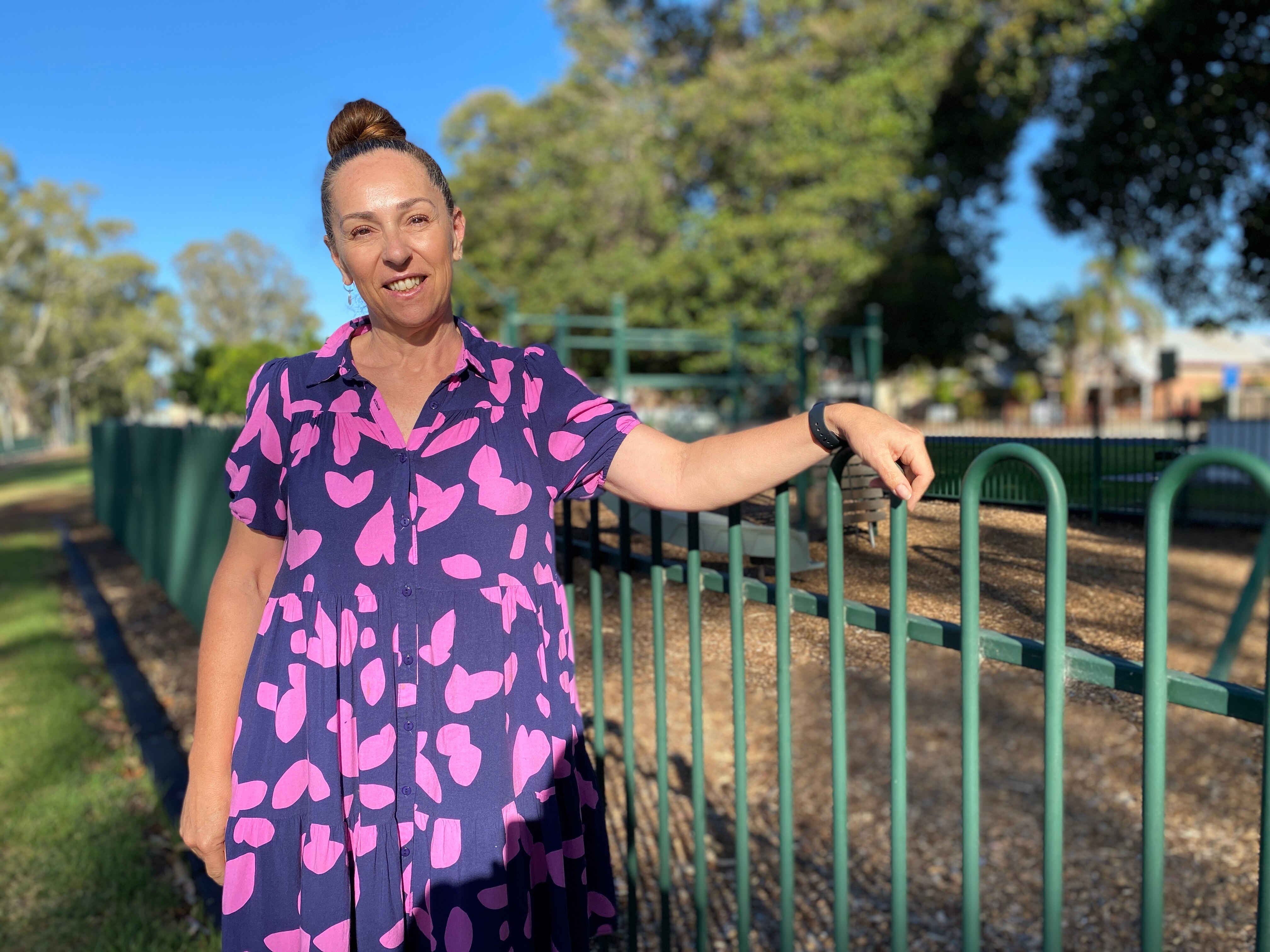A woman smiles with brown hair in a tight bun, she wears a navy and pink dress with trees behind