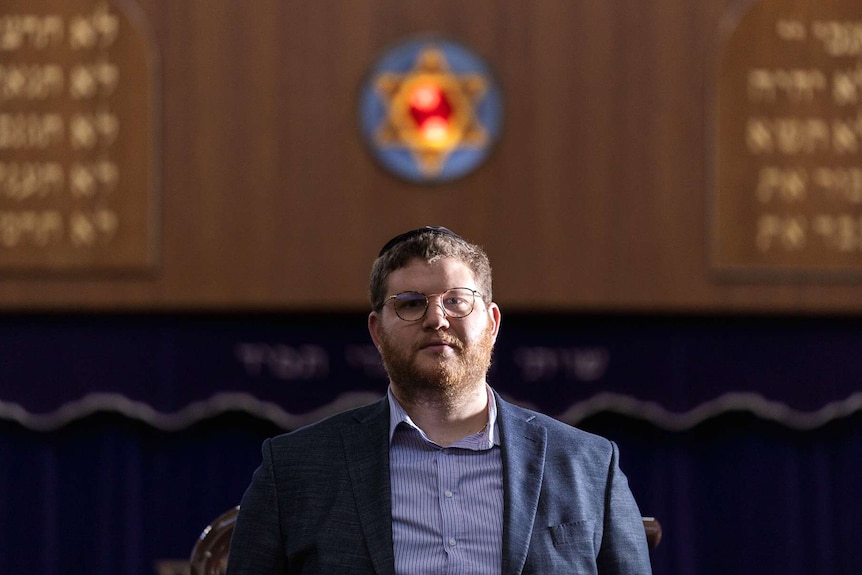 A head and shoulders shot of Perth Hebrew Congregation chief Rabbi Daniel Lieberman standing below a star of david on a wall.