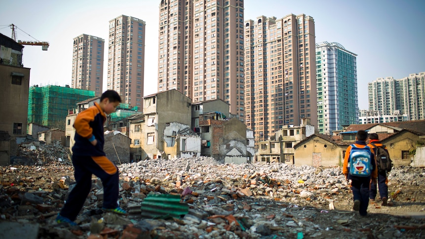 Three boys walk through rubble with a row of apartment towers along the skyline