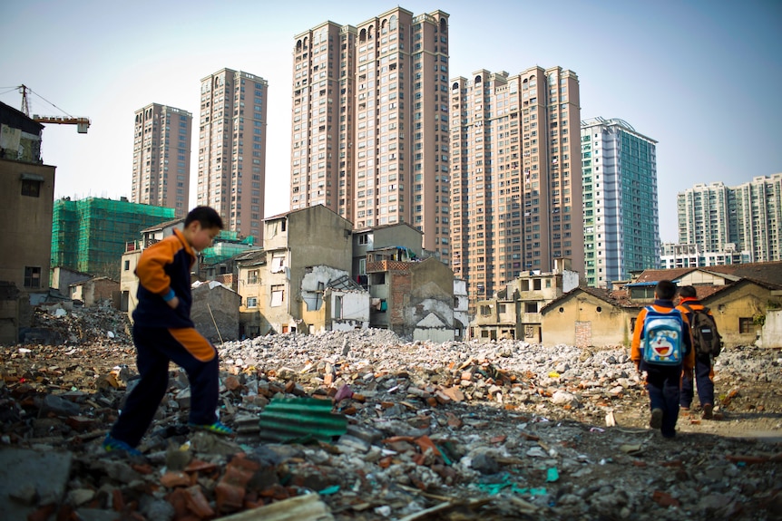 Three boys walk through rubble with a row of apartment towers along the skyline