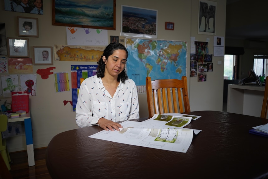 Rebecca sits at a wooden dining table in a white pattern shirt looking down at building plans.