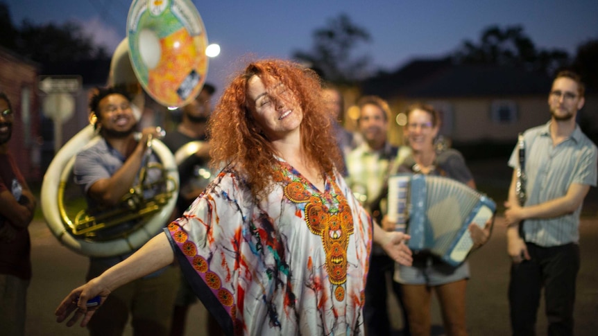 A woman with bright red curly hair is smiling with her eyes closed and arms spread out. A group of musicians are behind her.