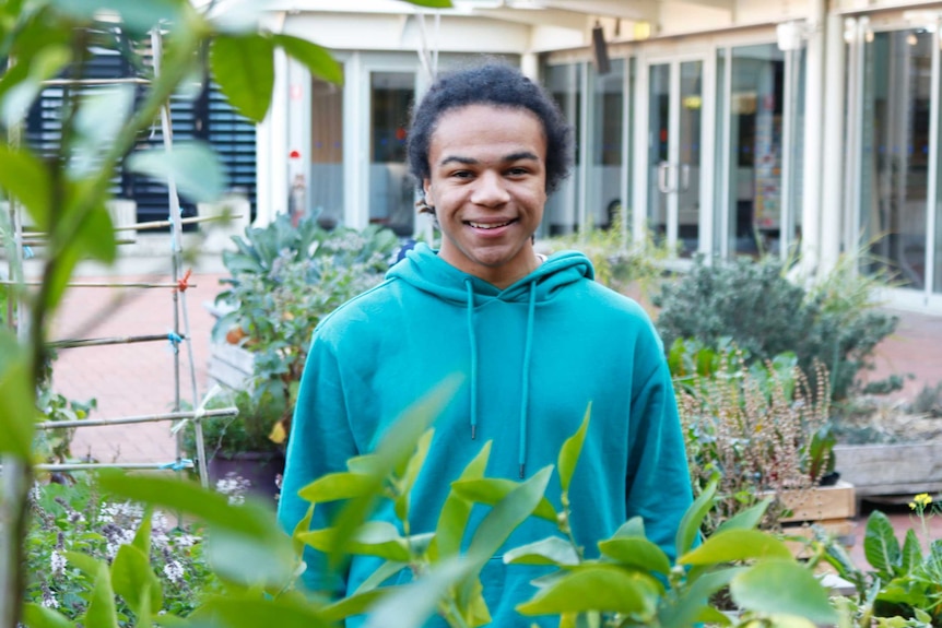 Young Indigenous man wearing a blue hoodie stands in a garden smiling into the camera. 