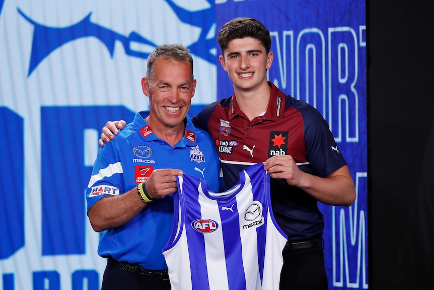 Harry Sheezel and Alastair Clarkson smile while standing together holding a North Melbourne jersey