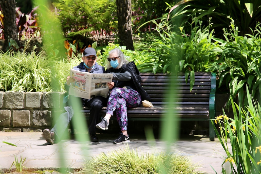 Two people in a park reading a newspaper sitting on a bench.