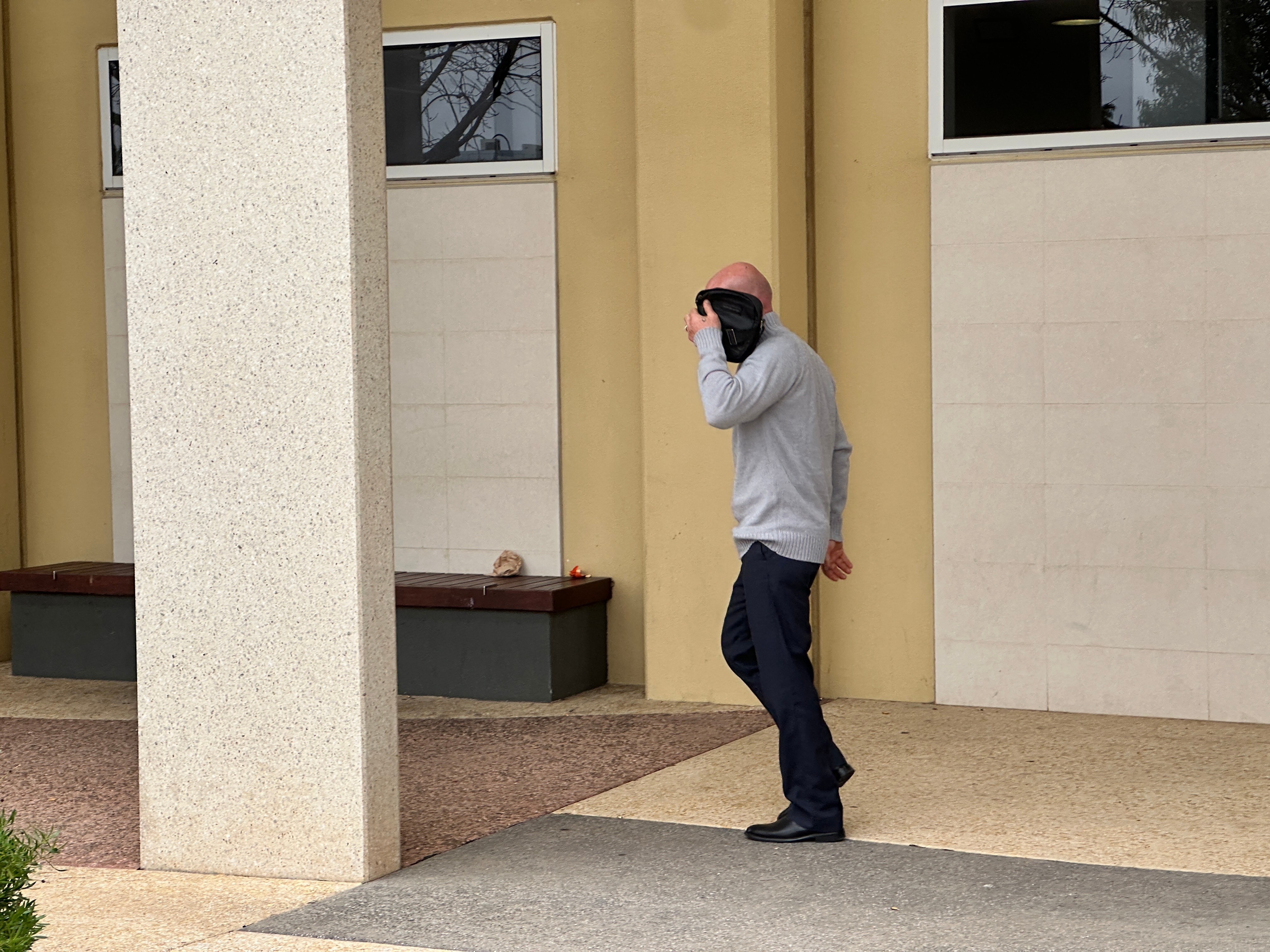 A man in a grey sweater and black pants uses a bag to cover his face