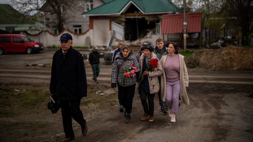 Three women link arms and carry flowers at front of a group along a dirty driveway. 