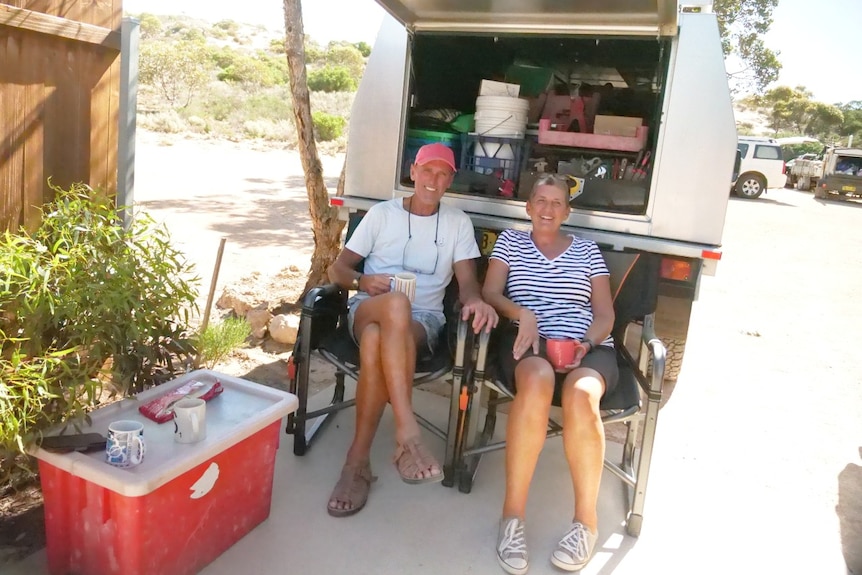 A man and a woman, sitting on fold-out chairs in front of a vehicle with an open cabin providing shade.